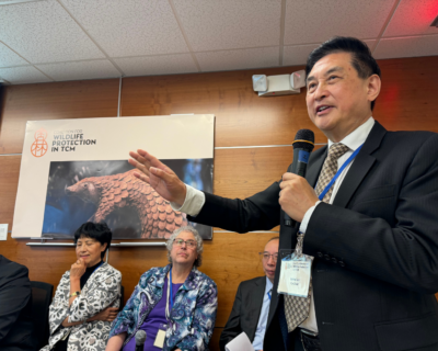 Dr. Lixing Lao holds a microphone as he addresses a room of people behind the camera. Four panelists sit behind him.